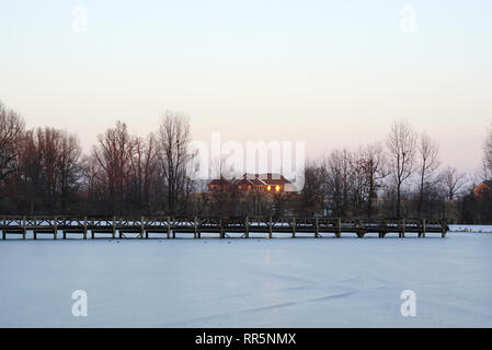 Coperta di neve park il marciapiede, lago di dock e case con fondo in lontananza al tramonto Foto Stock