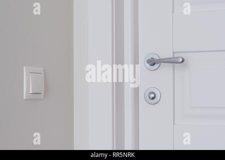 Dettaglio di un interno bianco porta con una delle porte cromate e maniglia di chiusura a scatto. Interruttore Beige sulla parete di luce sullo sfondo Foto Stock