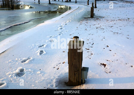 Coperta di neve park il marciapiede, lago di dock e case con fondo in lontananza al tramonto Foto Stock