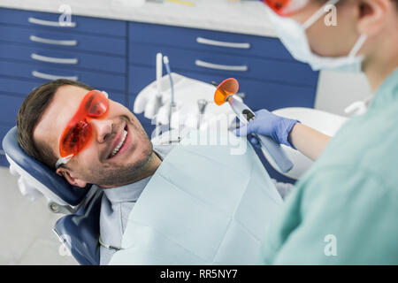 Messa a fuoco selettiva di allegro uomo in bicchieri vicino al dentista holding strumento dentale prima del procedimento di sbianca Foto Stock