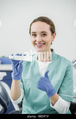 Allegro dentista femmina con rinforzi su denti tenendo il dentifricio e mostrando il pollice in alto Foto Stock