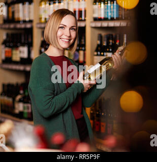 Immagine di donna felice con bottiglia nelle sue mani su sfondo sfocato di scaffali con bottiglie di vino in negozio Foto Stock
