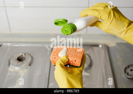 Due mani di applicare il sapone liquido dal flacone di detergente per spugna arancione. Estrusione di detersivo sulla spugna arancione su sfondo di fornelli a gas. Foto Stock