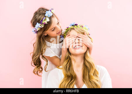 Carino bambino in piedi dietro la madre sorridente e chiude gli occhi con le mani su sfondo rosa Foto Stock