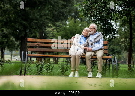 Sorridente coppia senior abbracciando e tenendo le mani mentre è appoggiato sul banco di legno in posizione di parcheggio Foto Stock