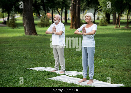 Felice coppia senior in piedi su Materassini da yoga in meditazione sukhasana pone in piedi con le mani giunte Foto Stock