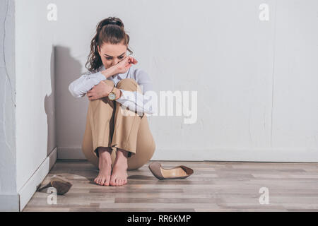 Triste donna in camicia bianca e pantaloni beige seduto sul pavimento e a piangere vicino alla parete di casa, addolorate concetto di disturbo Foto Stock