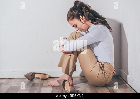 Triste donna in camicia bianca e pantaloni beige seduto sul pavimento con la testa piegata vicino al muro bianco a casa in lutto concetto di disturbo Foto Stock