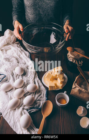 Giovane donna la cottura di una torta. Immagine ritagliata della donna seduta al tavolo azienda grande ciotola di vetro nei pressi di ingredienti per la cottura di torte fatte in casa. Foto Stock