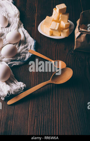 Ingredienti per la pasta frolla impasto marrone sul tavolo di legno, close-up. Piastra del burro a dadini, uova su asciugamano bianco e sacchetto di carta di farina accanto al rustico spo Foto Stock