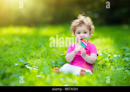 Baby girl a mangiare il gelato candy nel soleggiato parco. Bambino con lollipop. Bambini a giocare all'aperto in estate. Bambino con snack dolce. I bambini mangiano i dolci. Kid Foto Stock