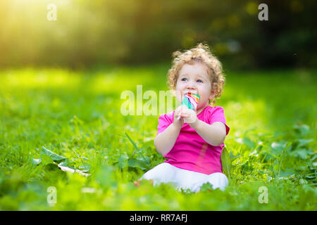 Baby girl a mangiare il gelato candy nel soleggiato parco. Bambino con lollipop. Bambini a giocare all'aperto in estate. Bambino con snack dolce. I bambini mangiano i dolci. Kid Foto Stock