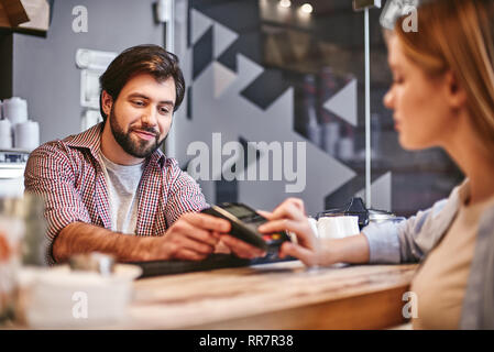 Sorridente dai capelli scuri serve barista donna in piedi vicino al bancone bar in cafe, il pagamento per l'ordine Foto Stock