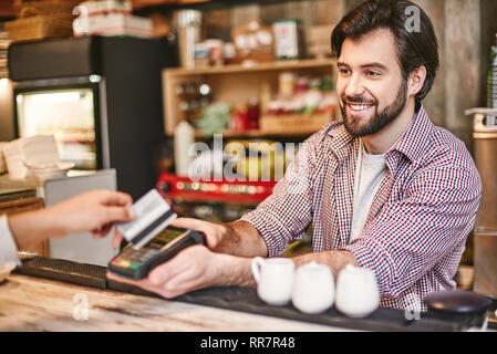 Sorridente dai capelli scuri serve barista donna in piedi vicino al bancone bar in cafe, il pagamento per l'ordine Foto Stock