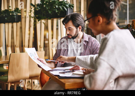 Due allegro colleghi per discutere di nuove idee seduto sulla scrivania. Barbuto uomo caucasico e attraente americano africano donna che lavorano con grafici. Telefono, documenti di lavoro e le matite sono sulla scrivania. Stile casual. Concetto di successo Foto Stock
