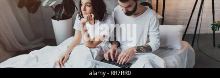 Uomo Barbuto sdraiato sul letto e utilizzando computer portatile mentre si annoia donna appoggiando il mento in camera da letto Foto Stock