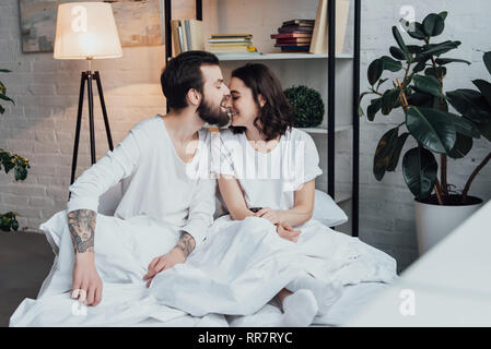 Bel uomo barbuto di mordere il naso della donna sorridente a casa nel letto Foto Stock