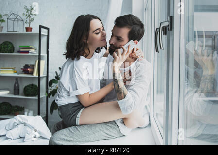 Bella donna che abbraccia l'uomo parlando sullo smartphone a casa Foto Stock