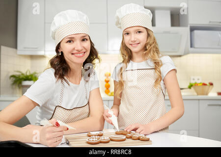 Splendida madre e carino daugter in grembiuli e cappelli chef seduti al tavolo. Giovane donna e bambina decorare gustosi biscotti di zucchero bianco smalto. American c Foto Stock