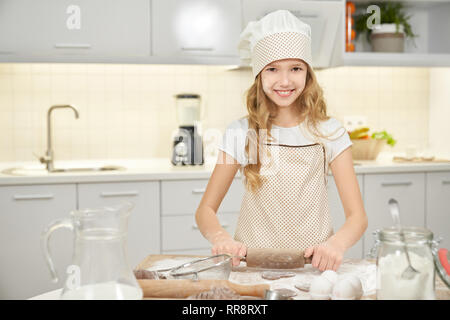 Affascinante adolescente permanente sulla cucina, tenendo matterello per la cottura dei biscotti. Bella ragazza che indossa in grembiule e chef hat guardando la telecamera, sorridente e Foto Stock