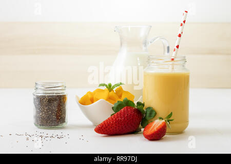 Bicchiere di mango fresco o smoothie mango frullato con il latte nella brocca di vetro, pezzi di mango in vaso di porcellana e qualche reale di fragole fresche a Pentecoste Foto Stock