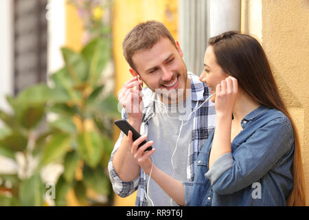 Coppia felice di condividere musica e di auricolari utilizzando uno smart phone in piedi in strada Foto Stock