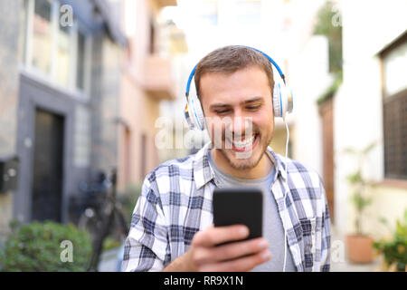 Vista frontale - Ritratto di un uomo felice ascoltando musica utilizzando smart phone a piedi in strada Foto Stock