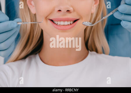 Vista ritagliata di dentista tenendo la bocca specchio e sonda dentale vicino a donna sorridente Foto Stock