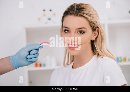 Bella donna sorridente guardando la fotocamera mentre il dentista di guanti in lattice che mantiene i denti coprire in clinica Foto Stock