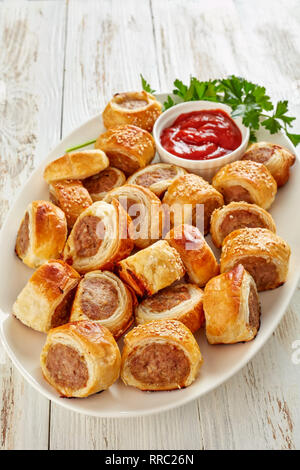 Pane appena sfornato di pasta sfoglia rotoli di salsiccia con ketchup e prezzemolo su una piastra bianca, partito inglese cibo, vista verticale dal di sopra, close-up Foto Stock