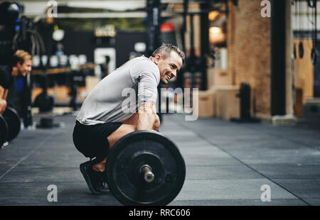 Montare l'uomo in sportswear sorridente durante la preparazione per il sollevamento pesi pesanti durante una sessione di allenamento in palestra Foto Stock