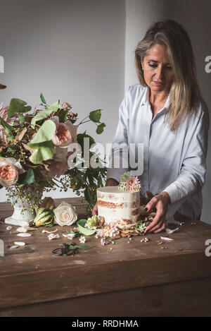 Vista laterale della signora posizionando il piatto con una gustosa torta decorata bloom bud sul tavolo di legno con mazzo di crisantemi, rose e rametti di piante in vaso tra Foto Stock