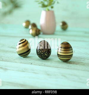 Tre di oro nero e uova di Pasqua decorate con strisce, splatter, fiori e macchie in oro nero e giallo su verde menta sullo sfondo di legno con copia Foto Stock