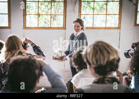 Vista posteriore dei cronisti riprese su telecamere donna felice che mostra il piatto con una gustosa torta decorata da bloom bud e foglie secche in camera Foto Stock