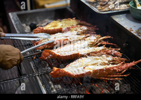 L uomo è la cottura di scampi alla griglia sulla strada del mercato alimentare a Bangkok in Tailandia. Egli utilizzando chrome pinze. Primo piano la foto in orizzontale. Foto Stock