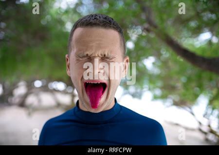 Ragazzo divertente con gli occhi chiusi e un grimace sta mostrando la sua lingua che dipinta in rosa luminoso sullo sfondo sfocato del tropicale sulla spiaggia di sabbia sulla Gi Foto Stock