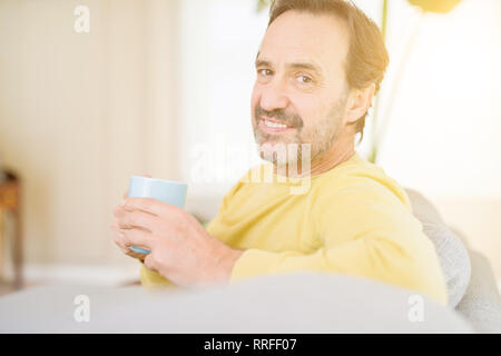 Bella la mezza età uomo seduto sul divano rilassante bevendo una tazza di caffè e sorridente con la videocamera a casa Foto Stock