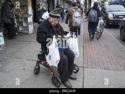 Uomo anziano si siede sul sedile del suo walker dopo lo shopping a riposo e verificare il suo telefono sul Kings Highway in Brooklyn, New York. Foto Stock