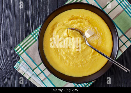 Hot polenta di mais con fondere il burro in una ciotola con il tovagliolo e cucchiaio d'argento su tavoli in legno nero, orizzontale vista da sopra Foto Stock