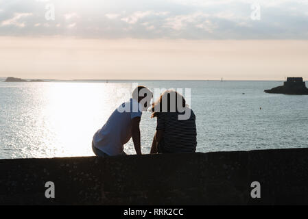 Vista posteriore del giovane ingannare intorno mentre è seduto sulla parete durante il tramonto contro ocean Foto Stock