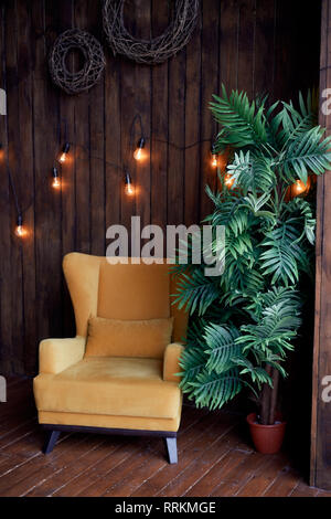 Accogliente centro senape-poltrona colorata, Palm tree e pareti di legno. Interno in stile retrò. Caldo giallo garland luce . Foto Stock