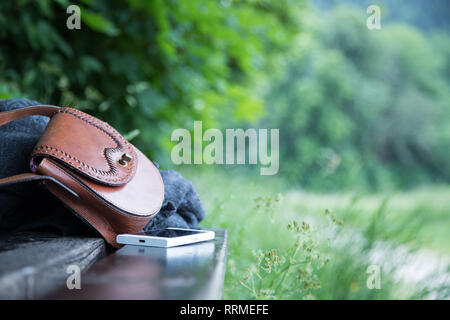 Pelle marrone borsetta, smartphone bianco e la camicia sono sdraiato su una panchina nel parco, nessuno, all'aperto Foto Stock