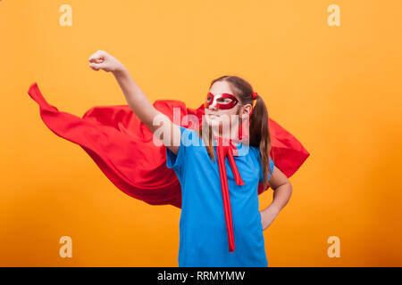 Little Girl in red cape gioca supereroe in studio su sfondo giallo. Infanzia felice. Fingendo di essere supereroe. Foto Stock