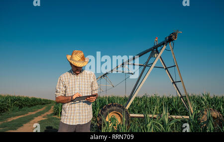 Grave agricoltore interessato utilizzando computer tablet in cornfield con impianto di irrigazione al di fuori del funzionamento durante le calde giornate estive Foto Stock