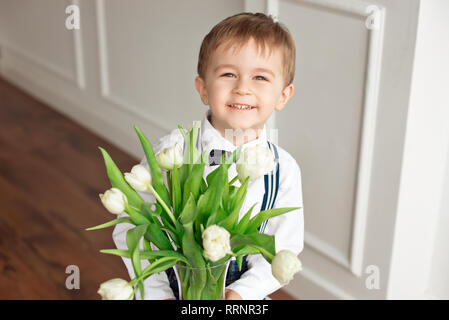 Carino ragazzo in una camicia bianca e un fiocco di Butterfly dà un mazzo di tulipani di colore bianco Foto Stock