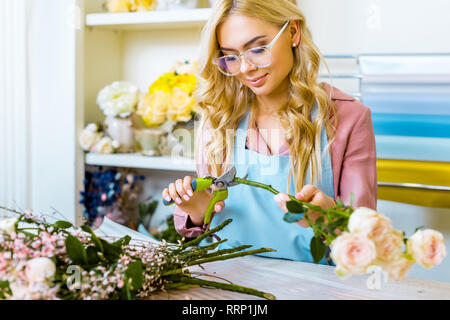 Messa a fuoco selettiva della bellissima femmina fioraio in bicchieri disponendo bouquet e il taglio di rose con potatore nel negozio di fiori Foto Stock