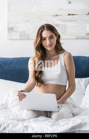 Bella donna incinta utilizzando computer portatile mentre è seduto sul letto Foto Stock