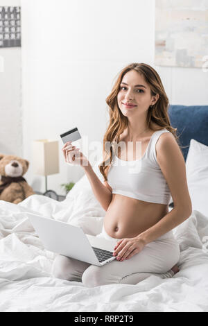 Felice incinta Donna seduta sul letto con il computer portatile e una carta di credito Foto Stock