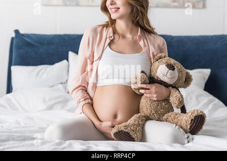 Vista parziale della donna incinta seduta sul letto e tenendo orsacchiotto Foto Stock