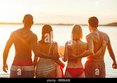 Specchietto di due diverse coppie giovani in costume da piedi a braccetto insieme in un lago di guardare il tramonto Foto Stock